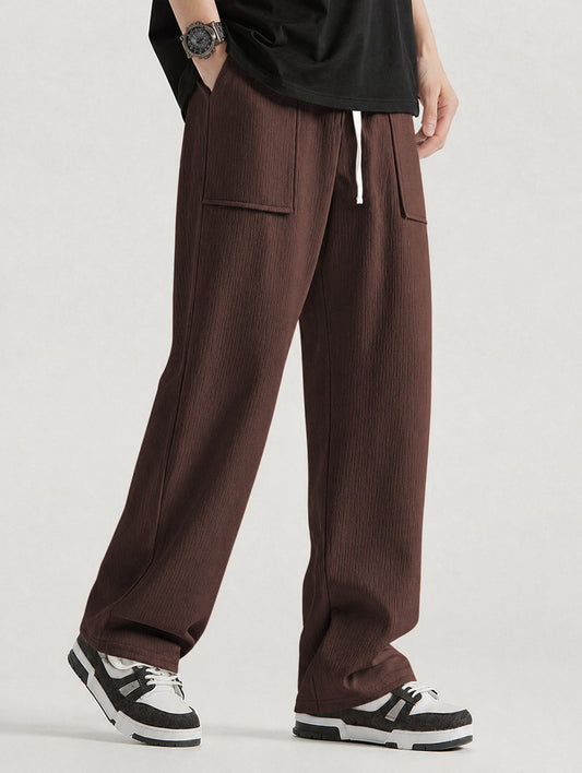 A person wearing loose, wide-leg coffee corduroy pants with a white drawstring, styled with a black t-shirt and black-and-white sneakers against a light gray studio background.