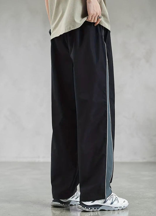 A person wearing loose black wide-leg track pants with a vertical gray side stripe, paired with a beige top and white sneakers, standing against a textured gray wall.
