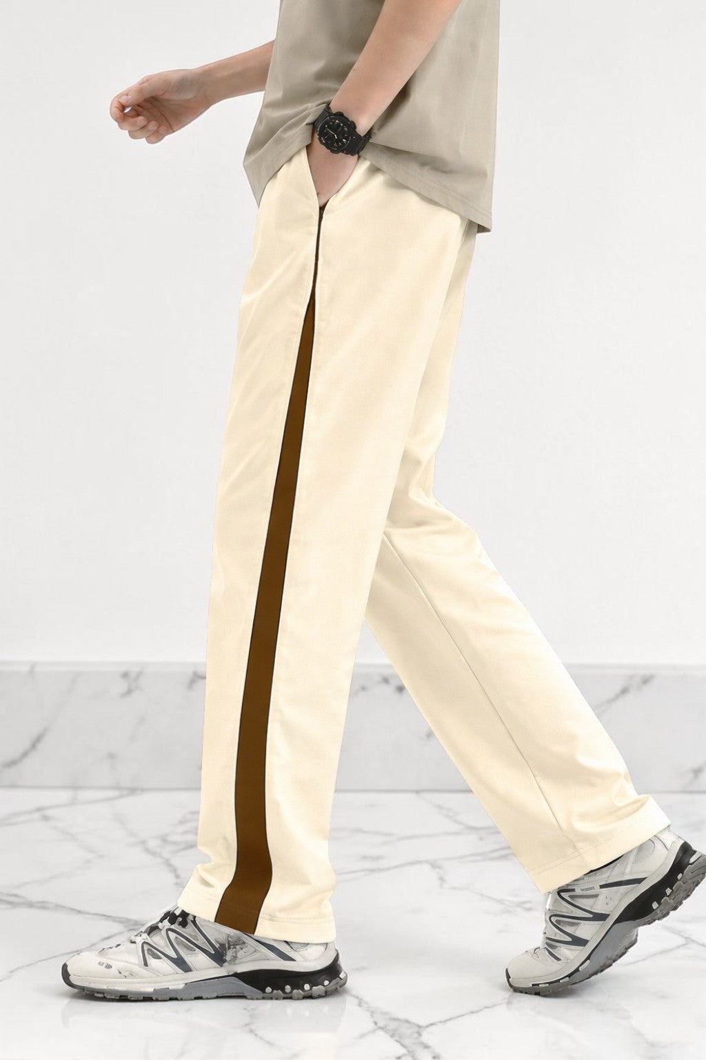 A person wearing cream-colored wide-leg track pants with a dark brown side stripe, paired with a beige t-shirt and gray sneakers, shown from the waist down against a white marble floor and wall.