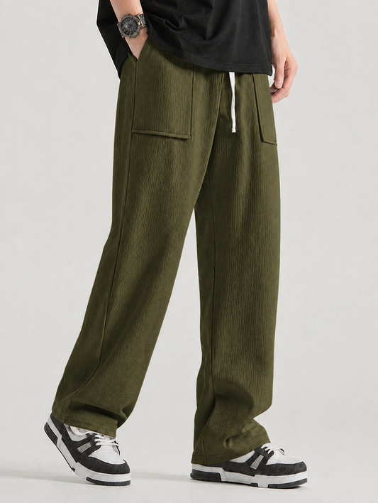 A person wearing loose olive green corduroy pants with side flap pockets, paired with a black t-shirt and black-and-white sneakers, shown from the waist down against a light gray studio background.