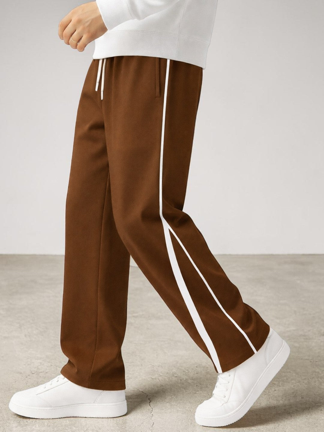 A person wearing coffee drawstring sweatpants with a cream side stripe, paired with white sneakers and a black backpack, shown from the waist down against a light studio background.