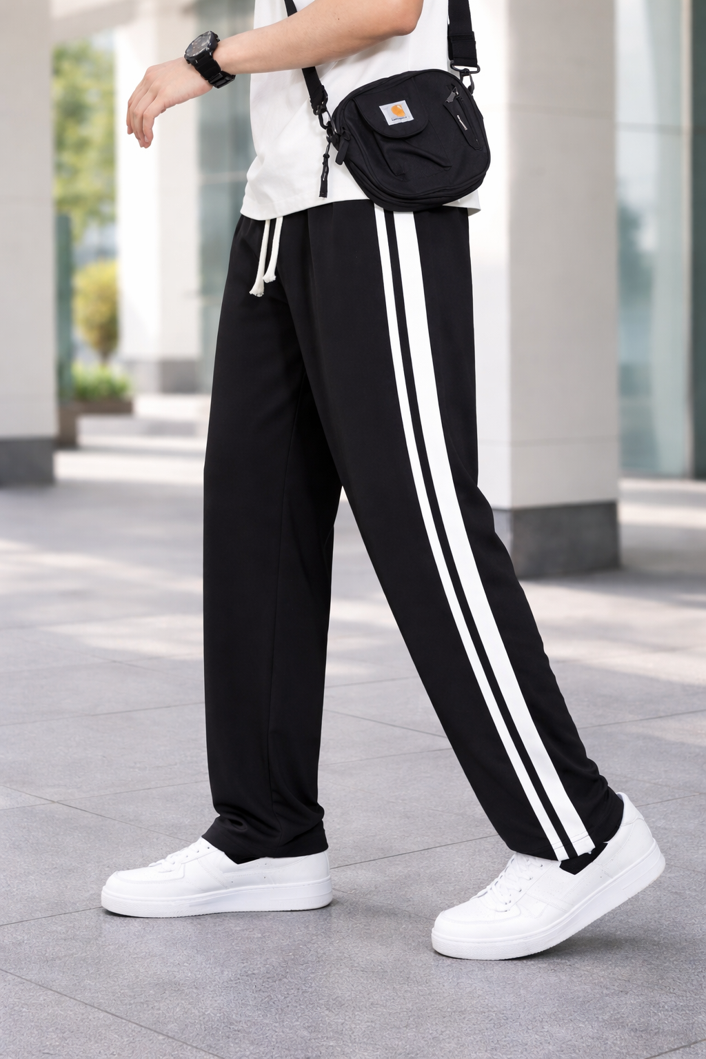 A person wearing black track pants with two white side stripes, white sneakers, and a black crossbody bag, standing outdoors on a paved walkway.
