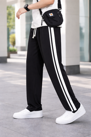 A person wearing black track pants with two white side stripes, white sneakers, and a black crossbody bag, standing outdoors on a paved walkway.