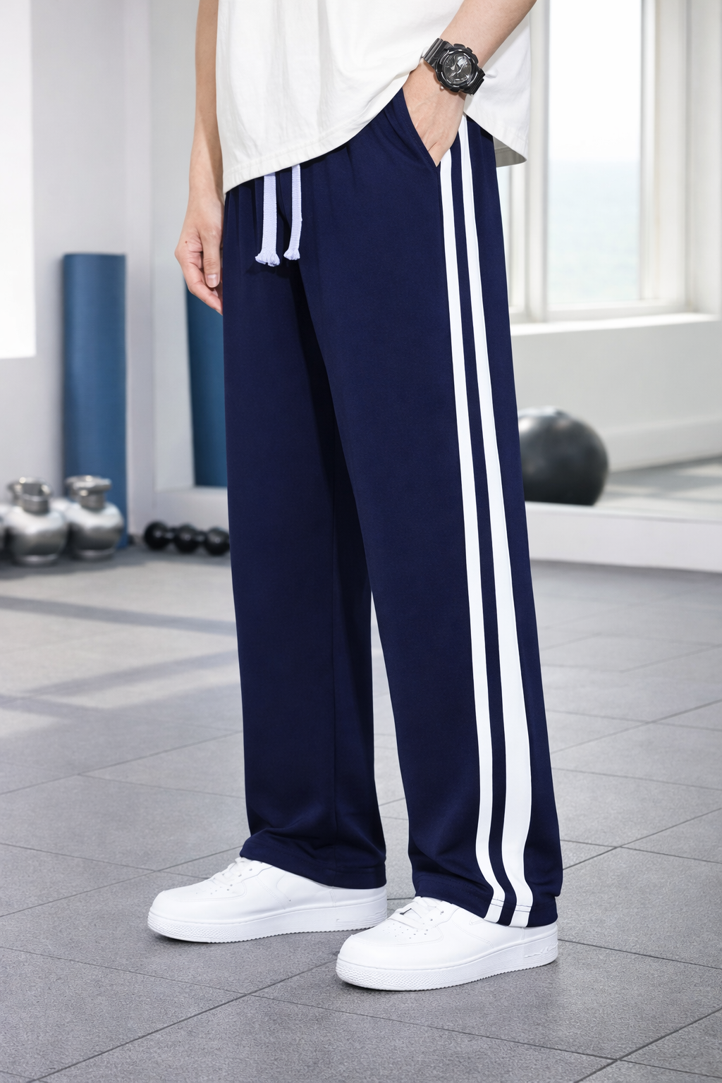 A person wearing navy blue athletic pants with two white side stripes, paired with a white t-shirt and white sneakers, standing in a gym setting.