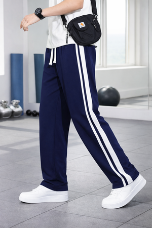 A person wearing navy blue athletic pants with two white side stripes, a white t-shirt, white sneakers, and a black crossbody bag, standing in a bright indoor space with exercise equipment in the background.
