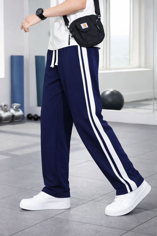 A person wearing navy blue athletic pants with two white side stripes, a white t-shirt, white sneakers, and a black crossbody bag, standing in a bright indoor space with exercise equipment in the background.