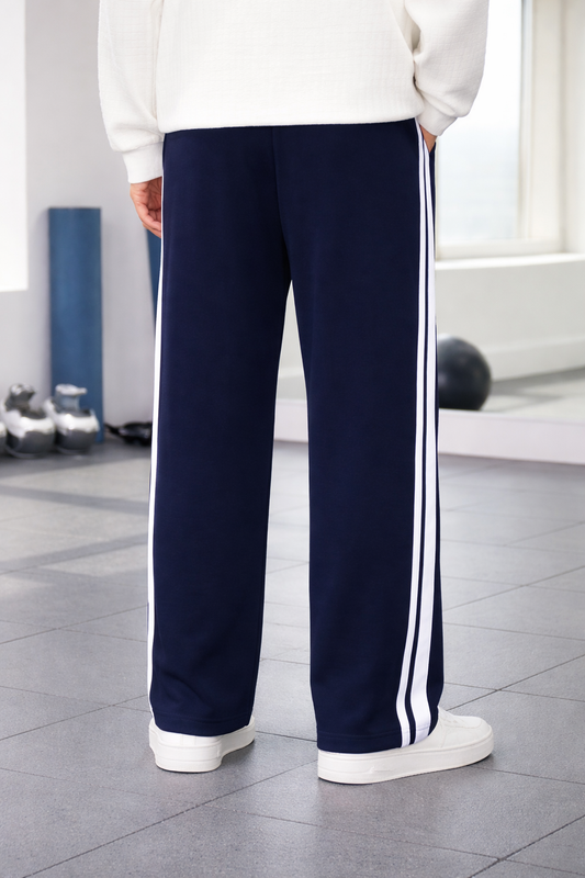 A person wearing navy blue wide-leg track pants with two white side stripes, paired with a white sweatshirt and white sneakers, standing in a gym.