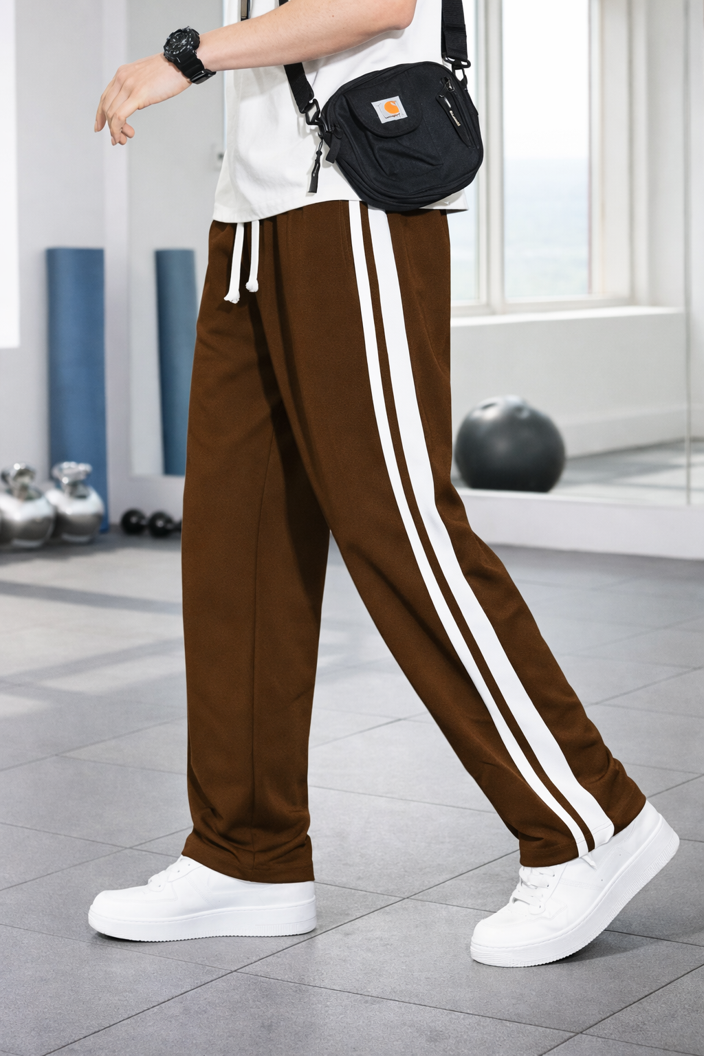 A person wearing coffee track pants with two white side stripes, a white t-shirt, white sneakers, and a black crossbody bag, standing in a gym setting.