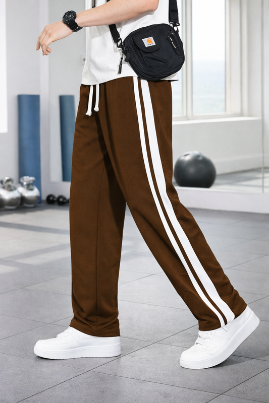 A person wearing coffee track pants with two white side stripes, a white t-shirt, white sneakers, and a black crossbody bag, standing in a gym setting.