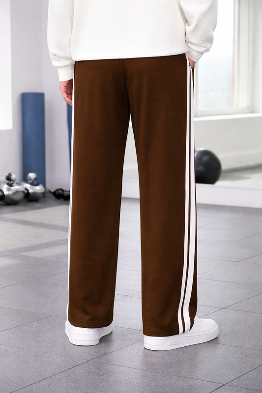 A person shown from the back wearing coffee wide-leg track pants with two white side stripes, a white sweater, and white sneakers in a bright indoor room with gym equipment in the background.