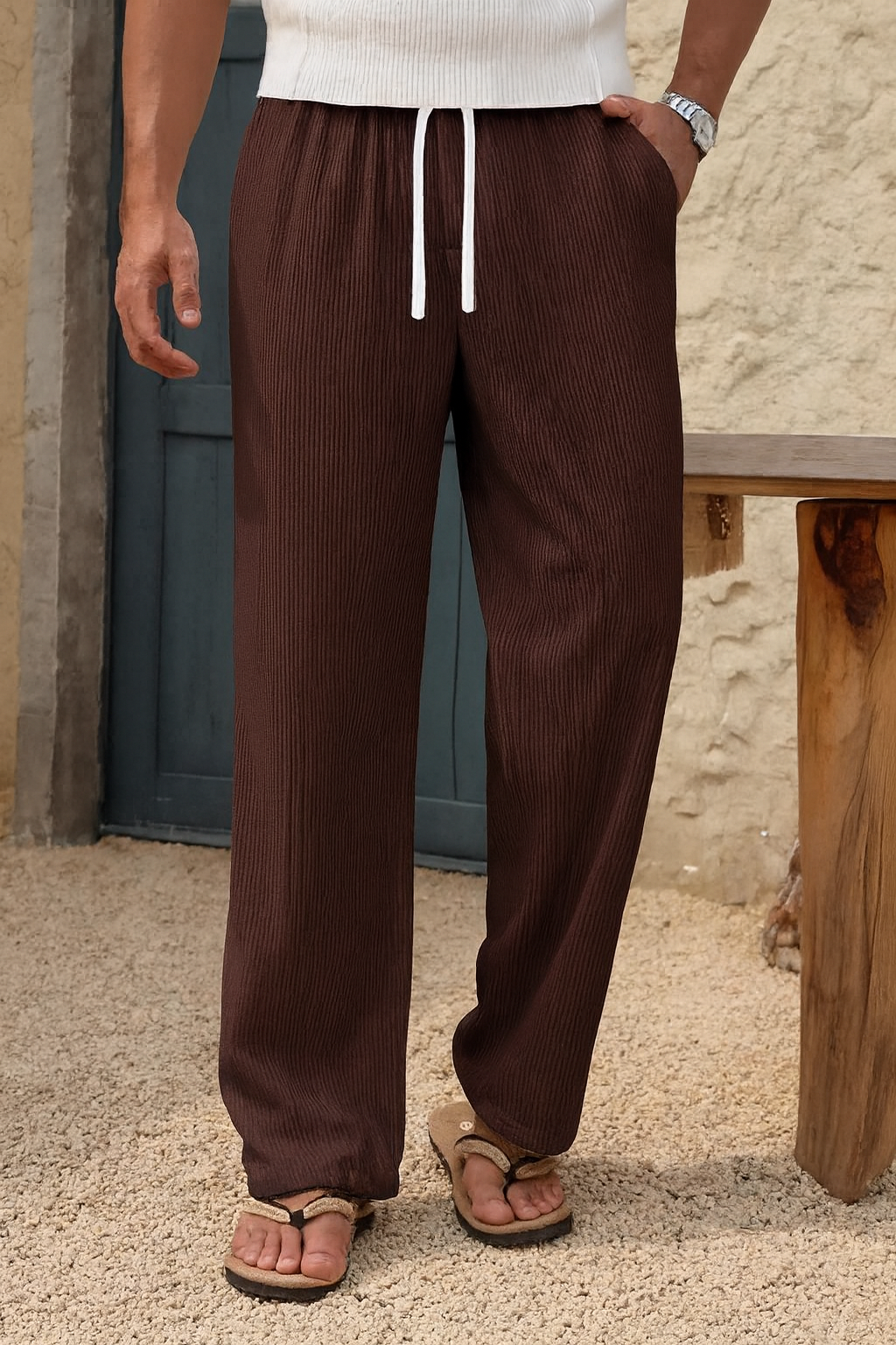 A person wearing coffee corduroy drawstring pants with a white drawstring, paired with a white ribbed top and tan sandals, standing outdoors next to a rustic wooden bench.