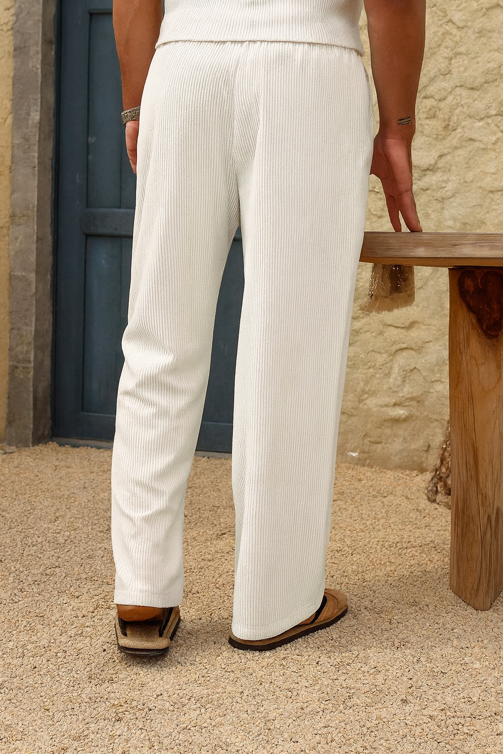 A person shown from the back wearing cream ribbed wide-leg trousers and brown sandals, standing on a textured stone floor next to a rustic wooden bench.