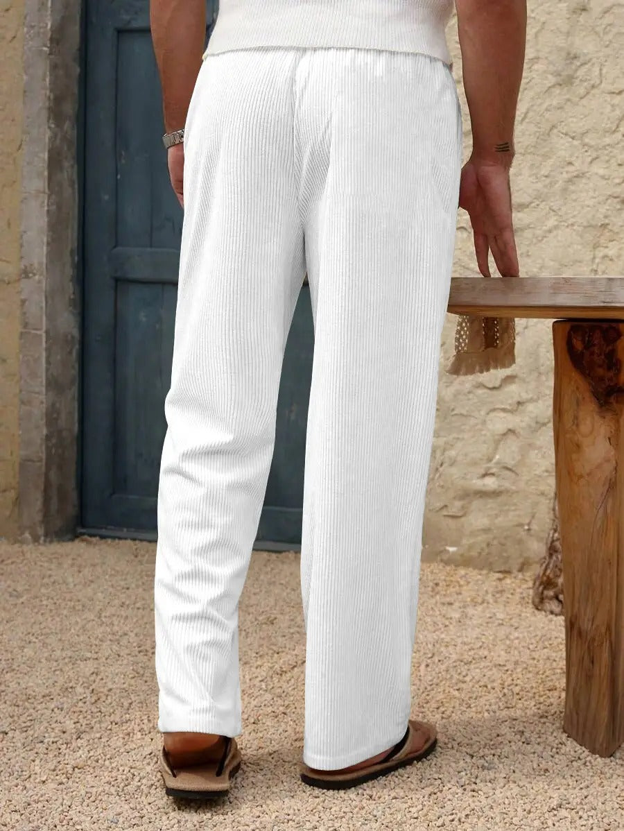 White ribbed knit wide-leg pants shown on a model from the back, paired with a white top and brown sandals, set against a rustic stone wall and wooden bench.