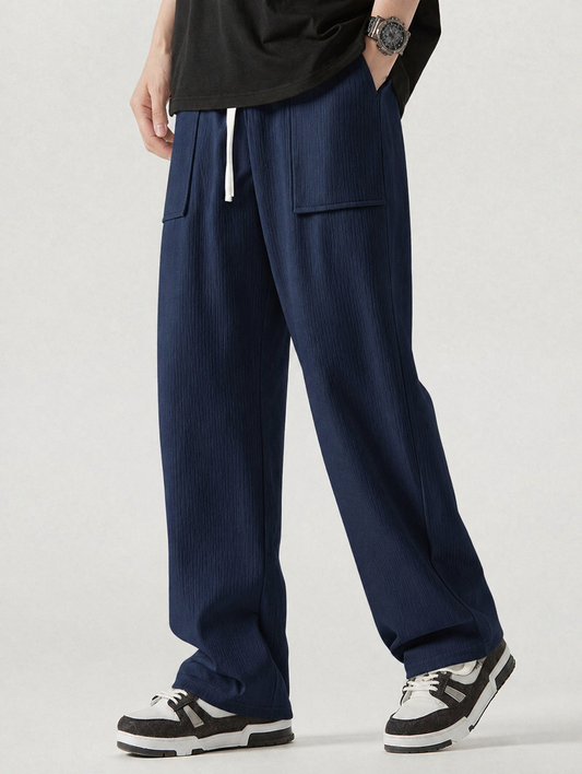 A person wearing loose, wide-leg navy pants with a white drawstring, shown from the waist down against a light gray studio background. The pants have a textured vertical rib pattern and large front patch pockets.