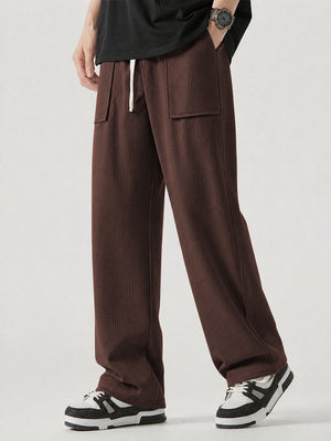A person wearing loose coffee corduroy pants with a white drawstring, paired with a black t-shirt and black-and-white sneakers, shown from the waist down against a light gray studio background.