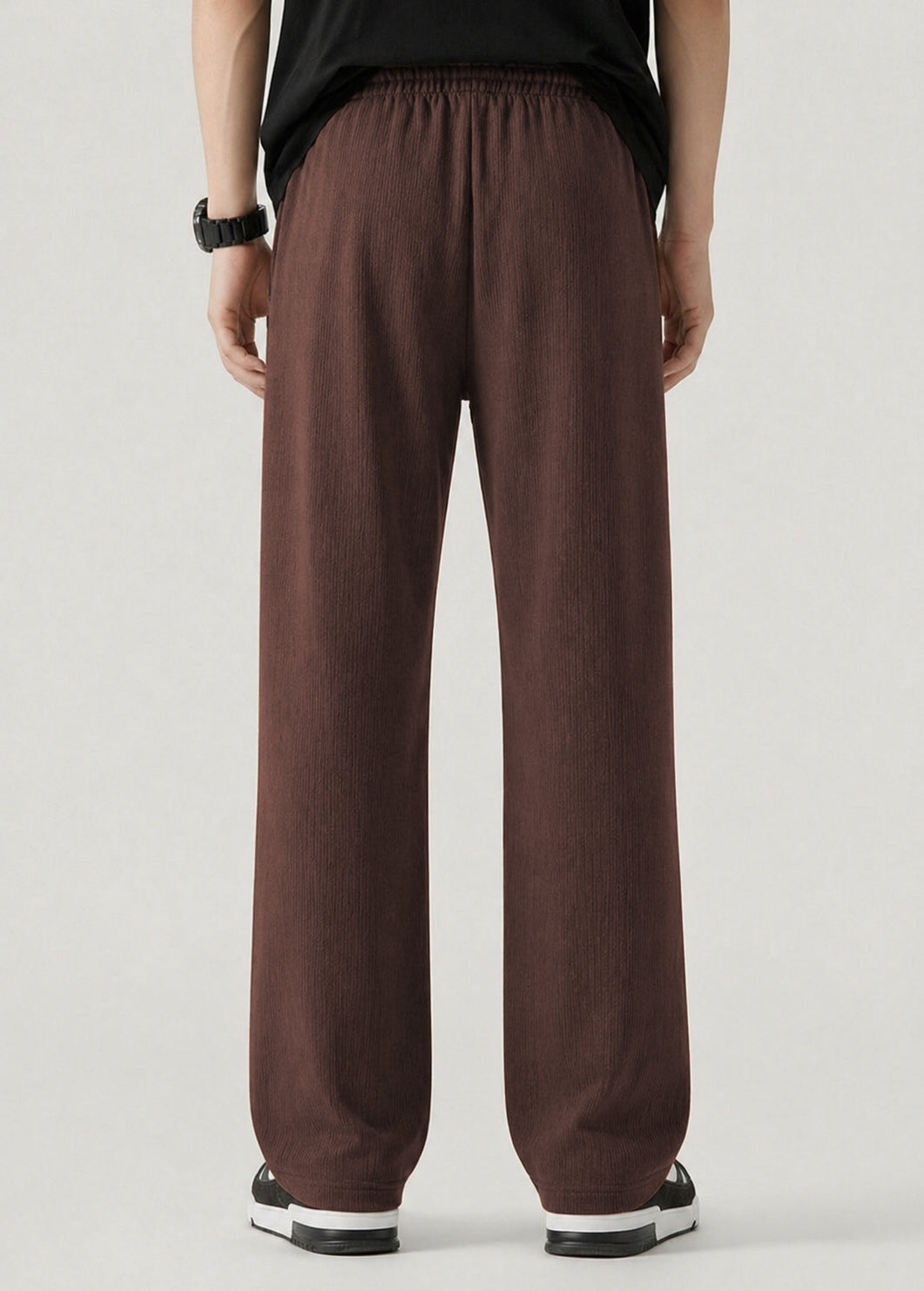 A person viewed from the back wearing loose, wide-leg coffee pants with an elastic waistband, paired with a black top and black-and-white sneakers, against a plain light gray studio background.
