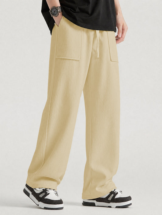 A person wearing wide-leg pleated trousers in a cream color, styled with a black t-shirt and black-and-white sneakers against a light gray studio background.