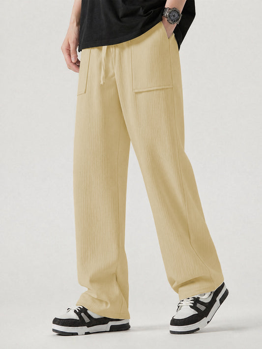 A person wearing loose, wide-leg pleated trousers in a cream, styled with a black t-shirt and black-and-white sneakers against a light gray studio background.