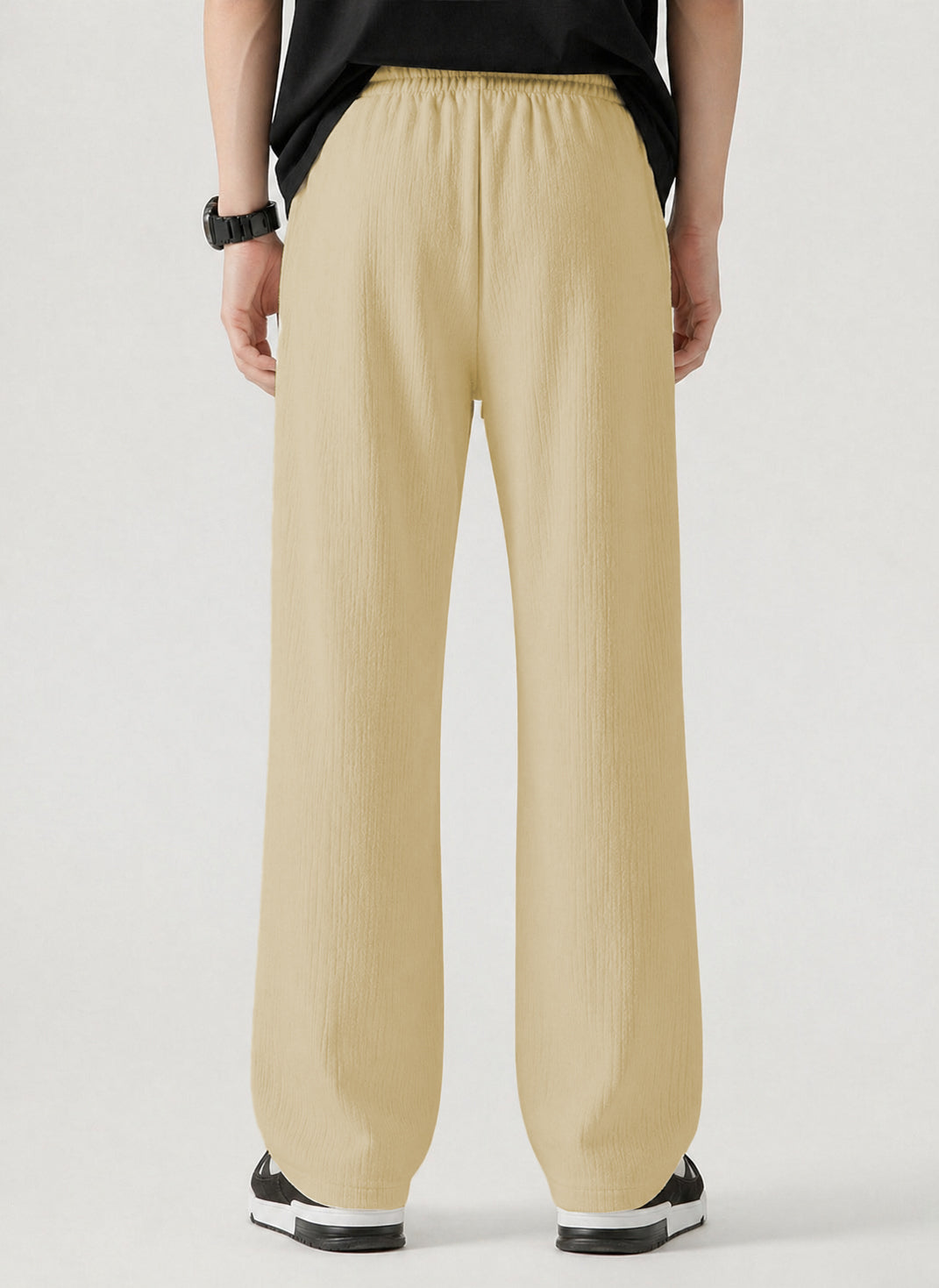 A person shown from the back wearing loose, wide-leg pants in a cream tone, paired with a black top and black-and-white sneakers, against a plain light gray studio background.