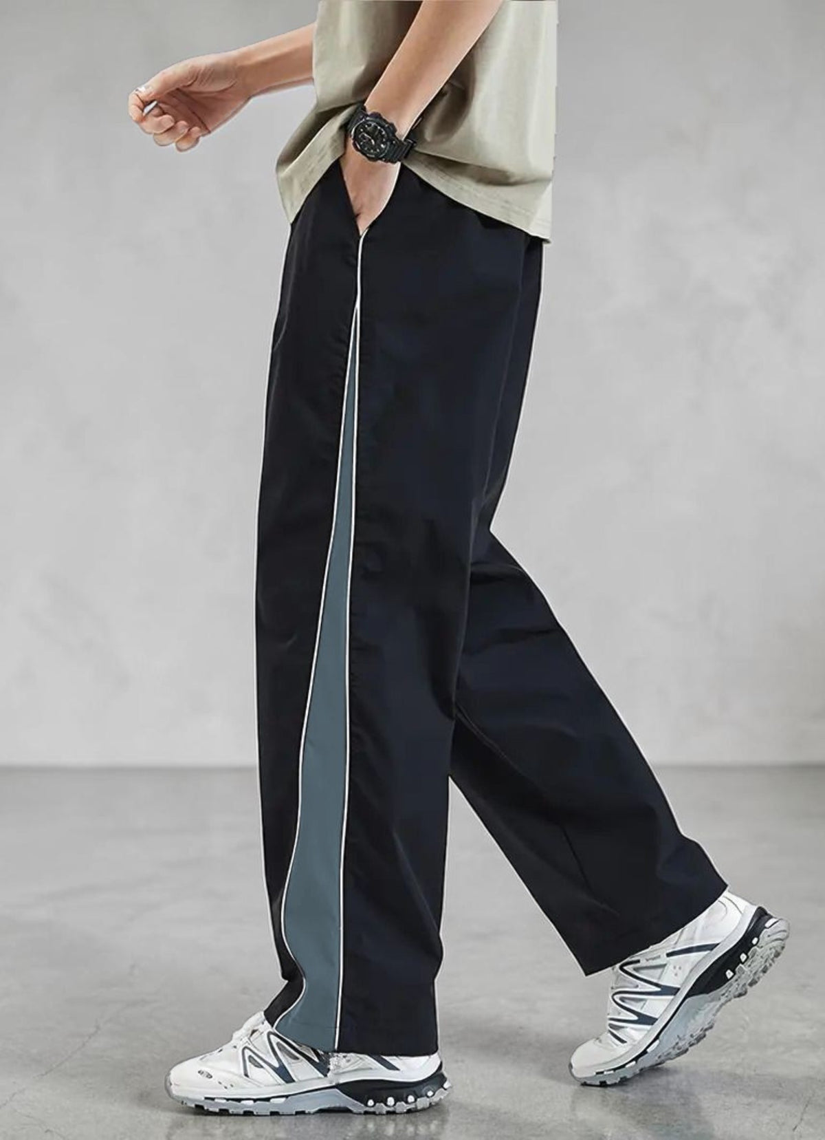 A person wearing loose black wide-leg pants with a vertical gray side stripe, paired with a light beige top and white sneakers, shown from the waist down against a gray studio background.
