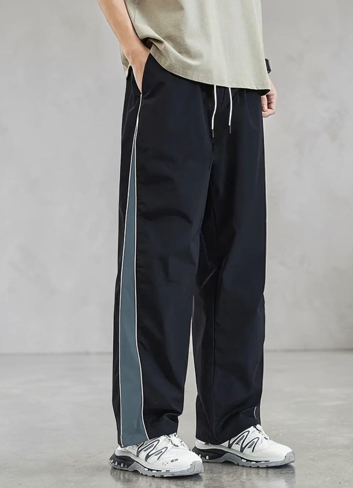 A person wearing loose black side-panel track pants with a gray stripe, a light beige t-shirt, and white sneakers, standing against a gray studio background.