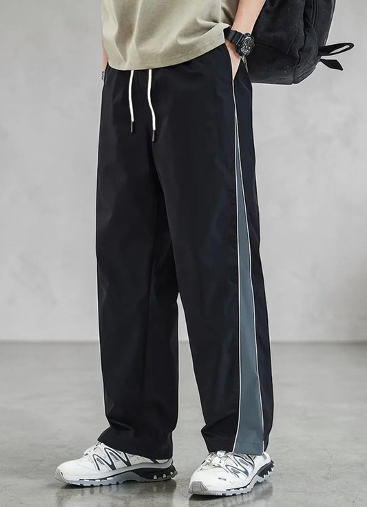 A person wearing loose black drawstring sweatpants with a vertical gray side stripe, paired with white sneakers and a black backpack, standing against a light gray studio wall.