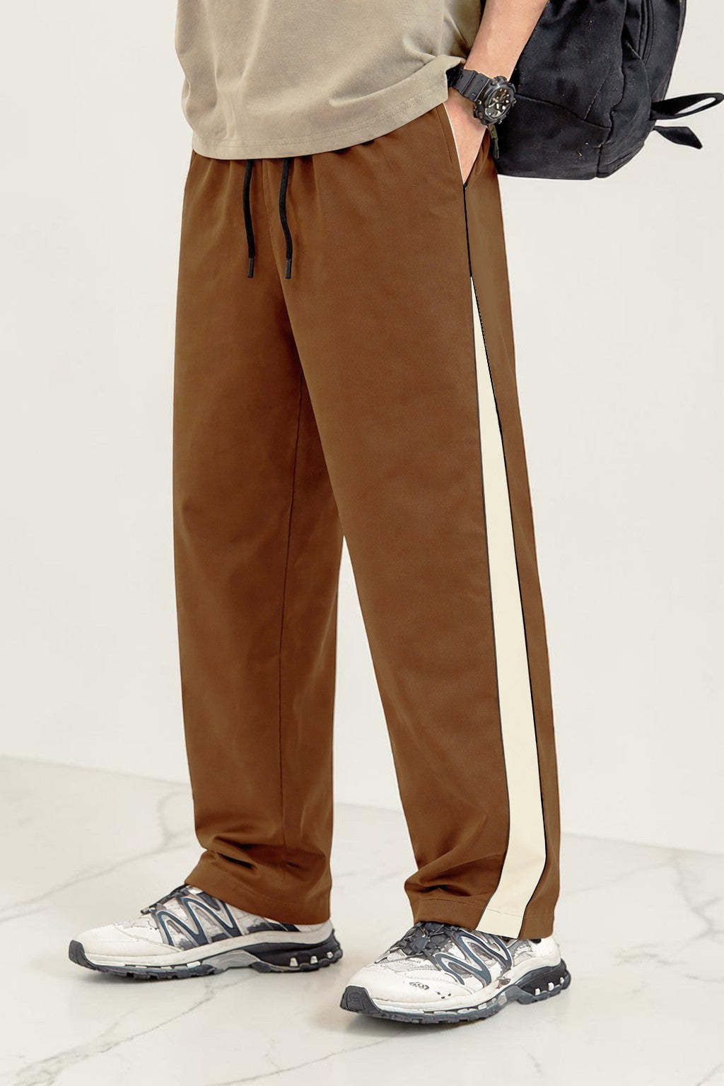 A person wearing coffee drawstring sweatpants with a cream side stripe, paired with white sneakers and a black backpack, shown from the waist down against a light studio background.