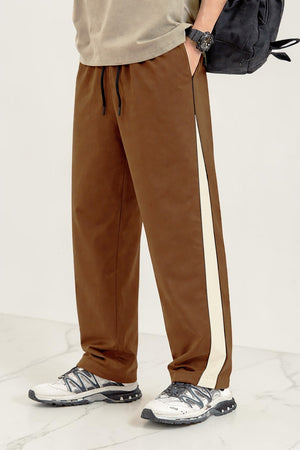 A person wearing coffee drawstring sweatpants with a cream side stripe, paired with white sneakers and a black backpack, shown from the waist down against a light studio background.