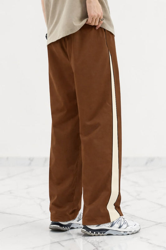 A person wearing coffee wide-leg track pants with a cream-colored side stripe, paired with a beige t-shirt and white sneakers, shown from the waist down on a light marble floor.