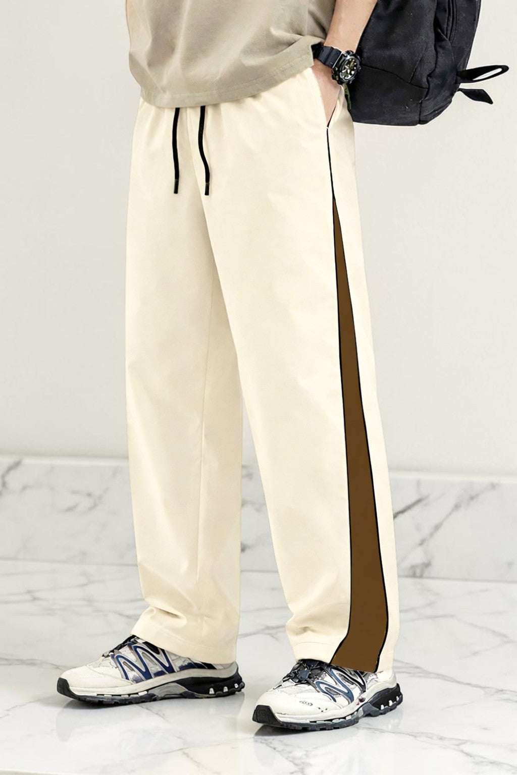 A person wearing cream-colored athletic pants with a dark brown side stripe, paired with white sneakers and a black backpack, shown from the waist down against a light gray marble background.