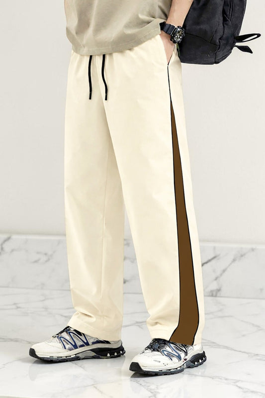 A person wearing cream-colored athletic pants with a dark brown side stripe, paired with white sneakers and a black backpack, shown from the waist down against a light gray marble background.