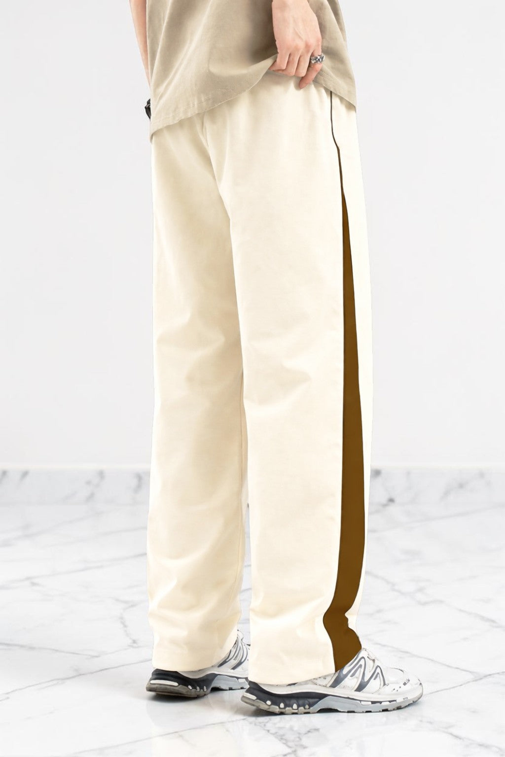 A person wearing cream wide-leg pants with a dark brown side stripe, paired with a beige top and white sneakers, standing on a light marble floor against a white wall.
