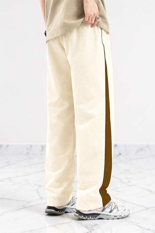 A person wearing cream wide-leg pants with a dark brown side stripe, paired with a beige top and white sneakers, standing on a light marble floor against a white wall.