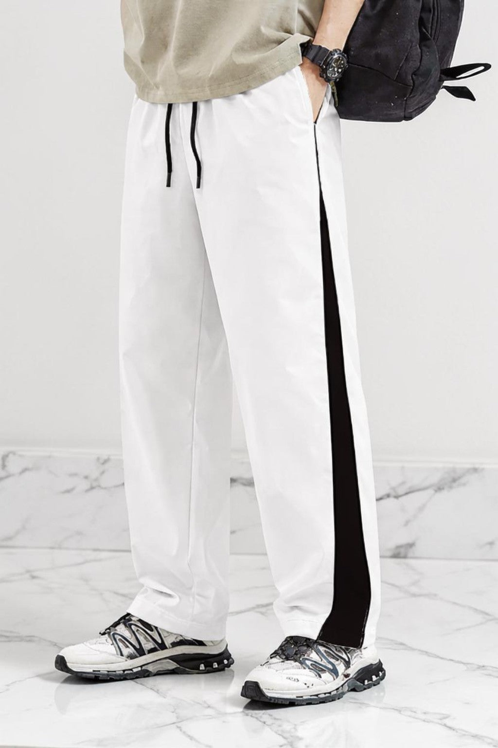 White athletic jogger pants with a black side stripe and black drawstrings, worn by a model with white and black sneakers, a beige top, and a black backpack against a marble floor and wall.