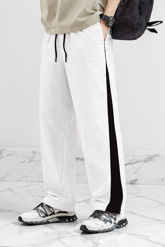 White athletic jogger pants with a black side stripe and black drawstrings, worn by a model with white and black sneakers, a beige top, and a black backpack against a marble floor and wall.