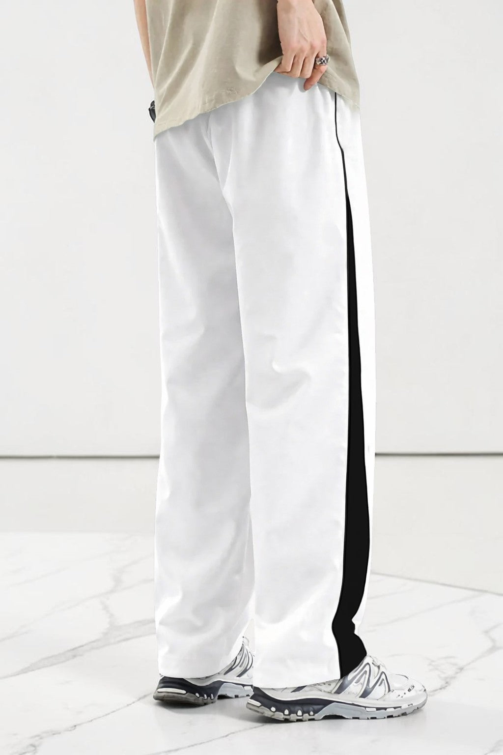 White wide-leg track pants with a bold black side stripe, modeled with a beige t-shirt and white sneakers on a light marble floor.