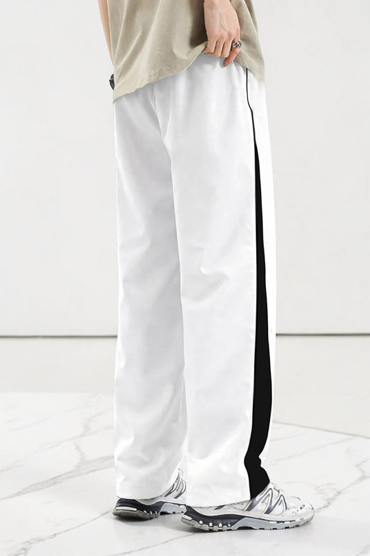 White wide-leg track pants with a bold black side stripe, modeled with a beige t-shirt and white sneakers on a light marble floor.