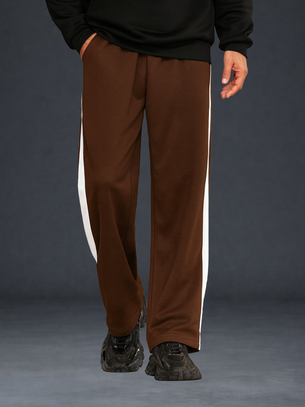 A person wearing coffee wide-leg track pants with cream side stripes, paired with a black sweatshirt and black sneakers, shown from the waist down against a dark gray studio background.