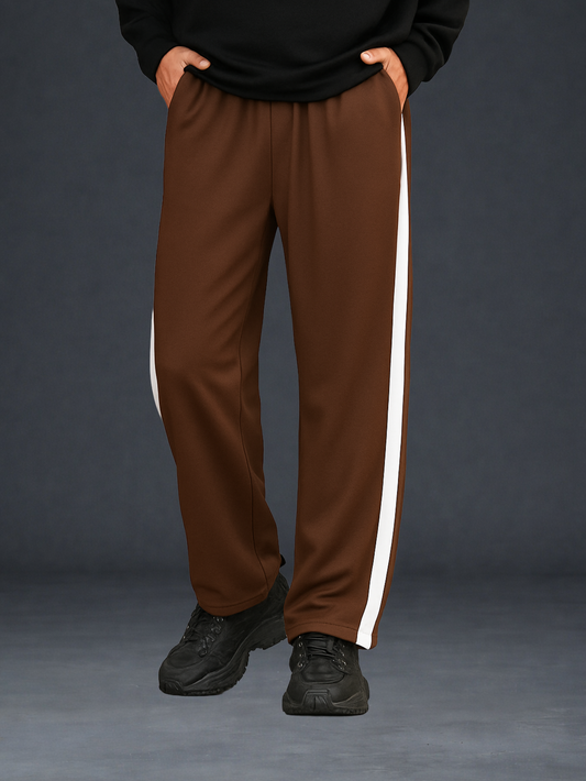 A person wearing coffee track pants with a cream side stripe, paired with a black sweatshirt and black sneakers, standing against a dark gray studio background.