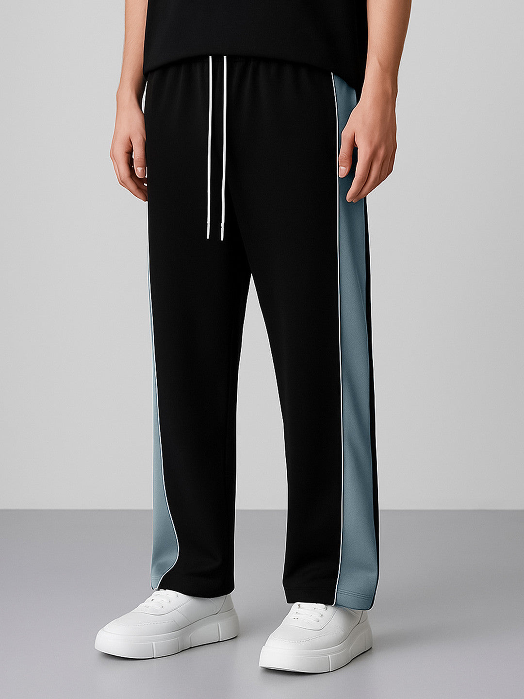 Black track pants with white drawstrings and light blue side stripes, worn by a model with white sneakers against a gray studio background.