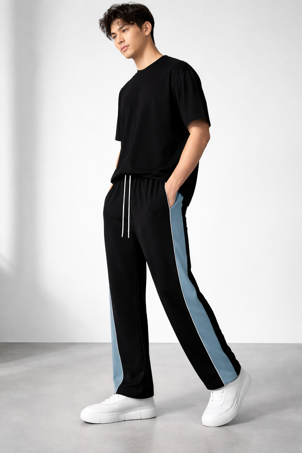 A man modeling black athletic pants with light blue side panels and white drawstrings, paired with a black t-shirt and white sneakers, in a studio setting.