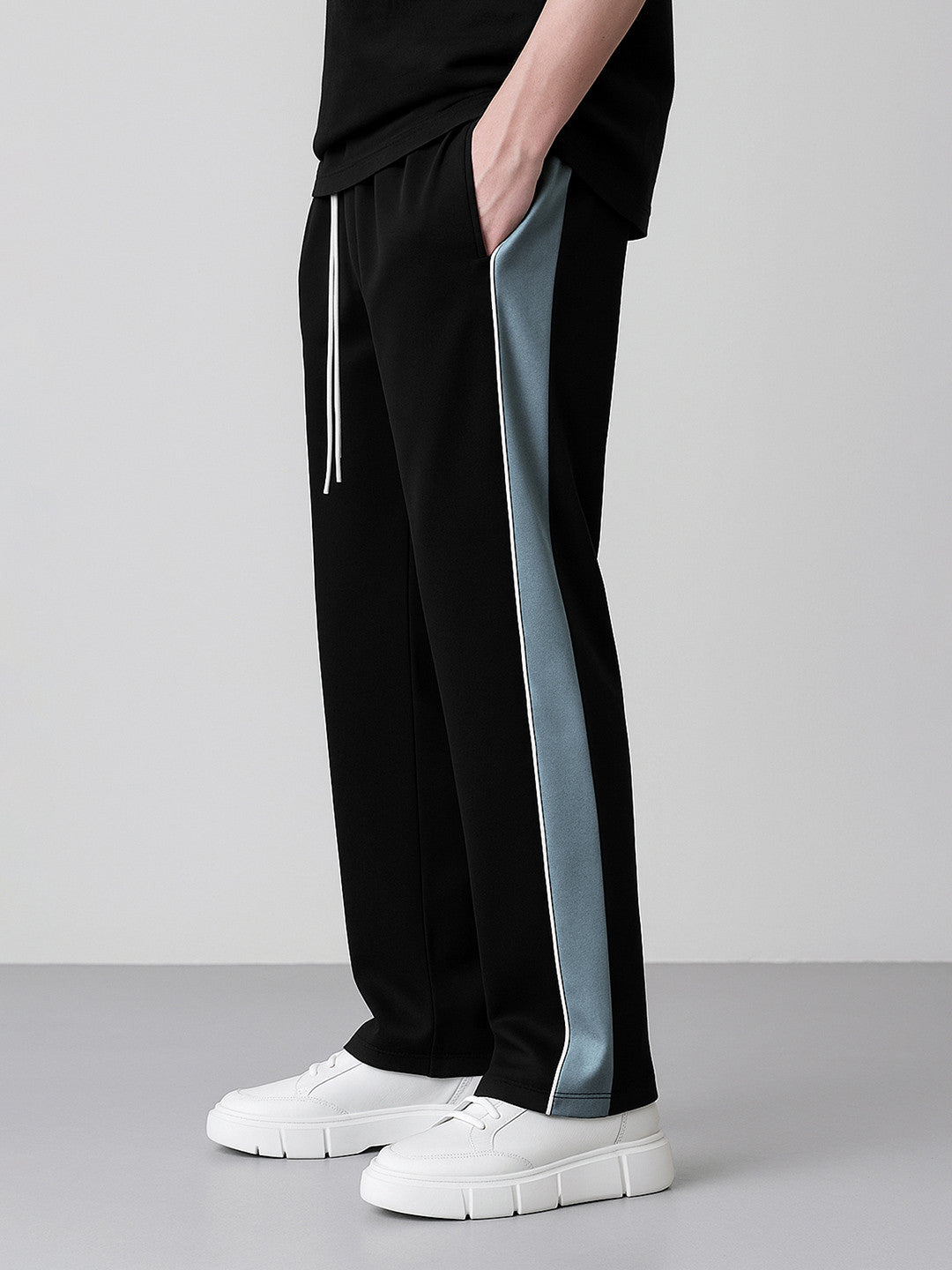 Black wide-leg track pants with a light blue side stripe and white drawstring, shown on a model wearing white platform sneakers against a neutral gray studio background.
