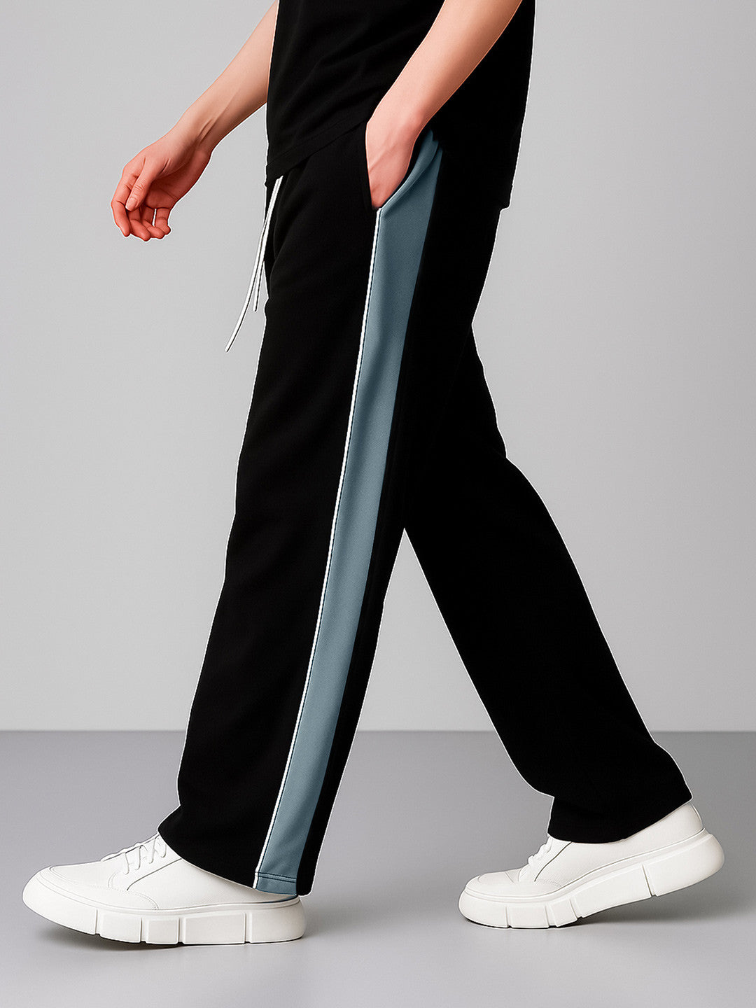 A person wearing black wide-leg sweatpants with a light blue-gray vertical stripe down the side, paired with white platform sneakers, shown against a neutral gray studio background.