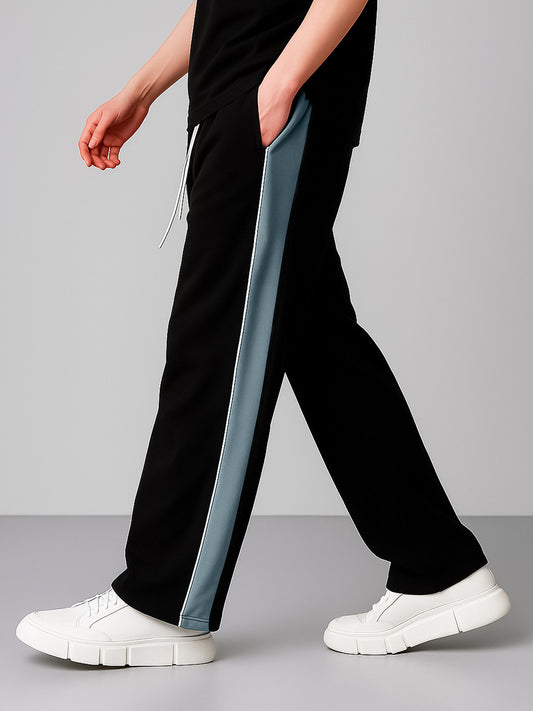 A person wearing black wide-leg sweatpants with a light blue-gray vertical stripe down the side, paired with white platform sneakers, shown against a neutral gray studio background.