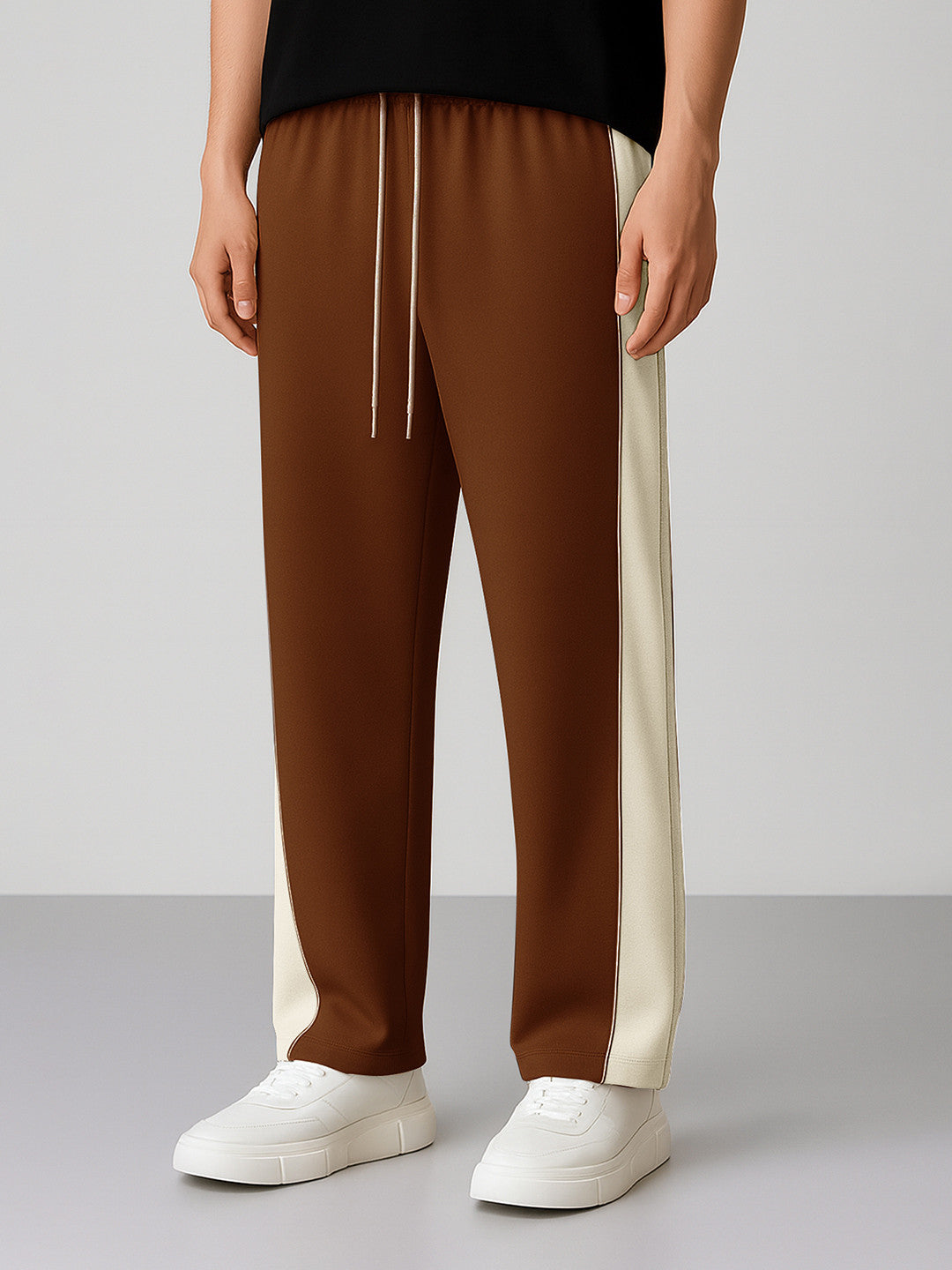 A person wearing coffee track pants with cream side panels and white drawstrings, paired with white sneakers, shown from the waist down against a light gray studio background.
