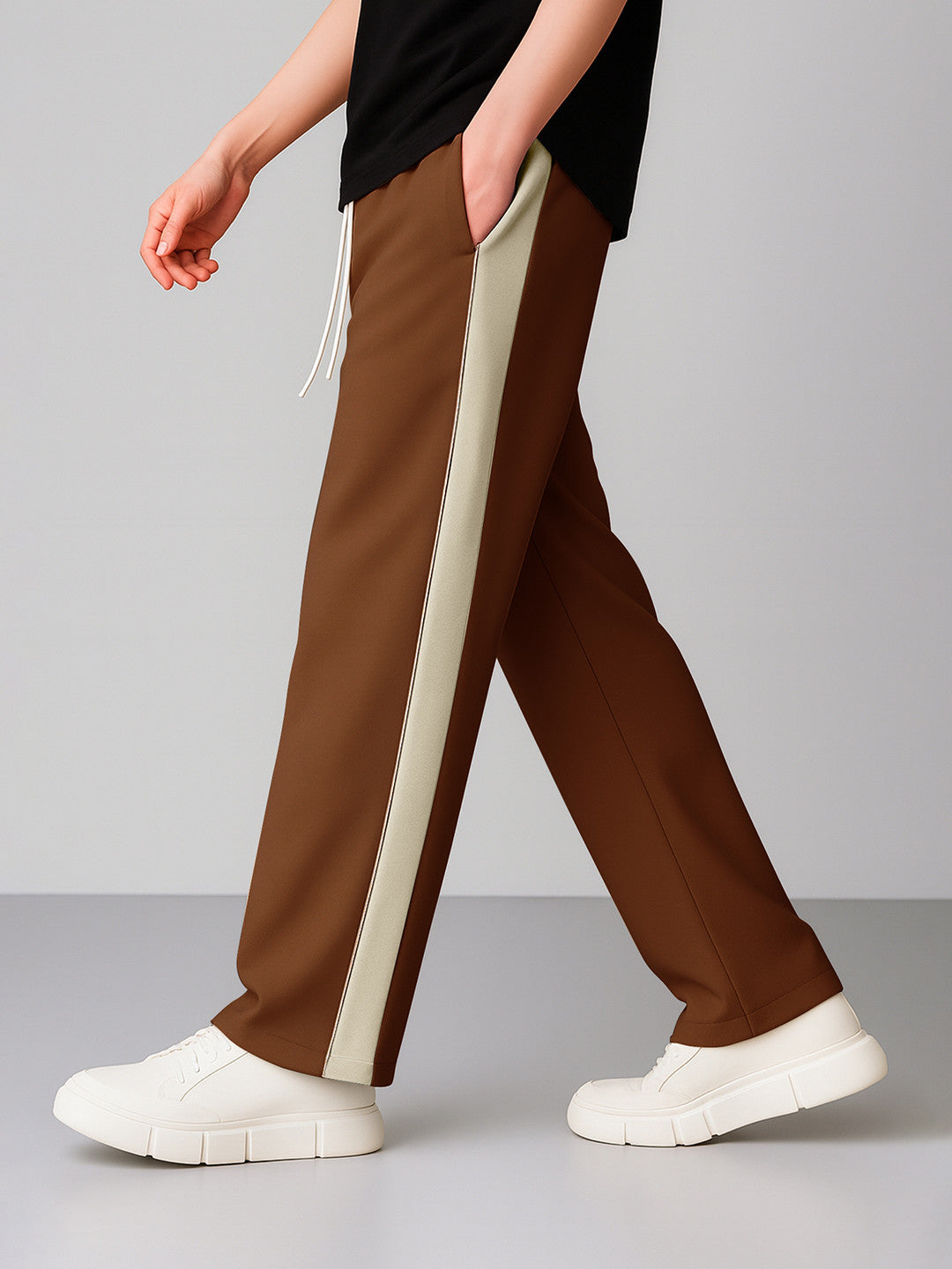 A person wearing coffee wide-leg pants with a wide cream side stripe, paired with a black top and white platform sneakers, shown from the waist down against a light gray studio background.