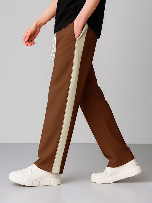 A person wearing coffee wide-leg pants with a wide cream side stripe, paired with a black top and white platform sneakers, shown from the waist down against a light gray studio background.