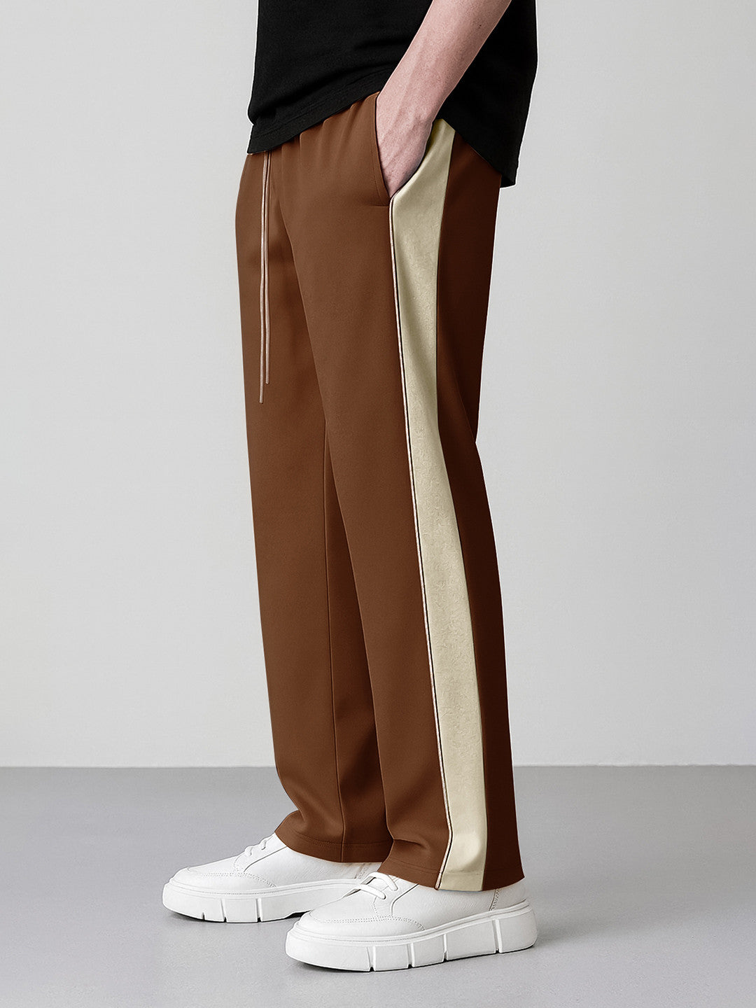 A person wearing coffee wide-leg pants with a beige side stripe, paired with a black top and white sneakers, shown from the waist down against a light gray studio background.