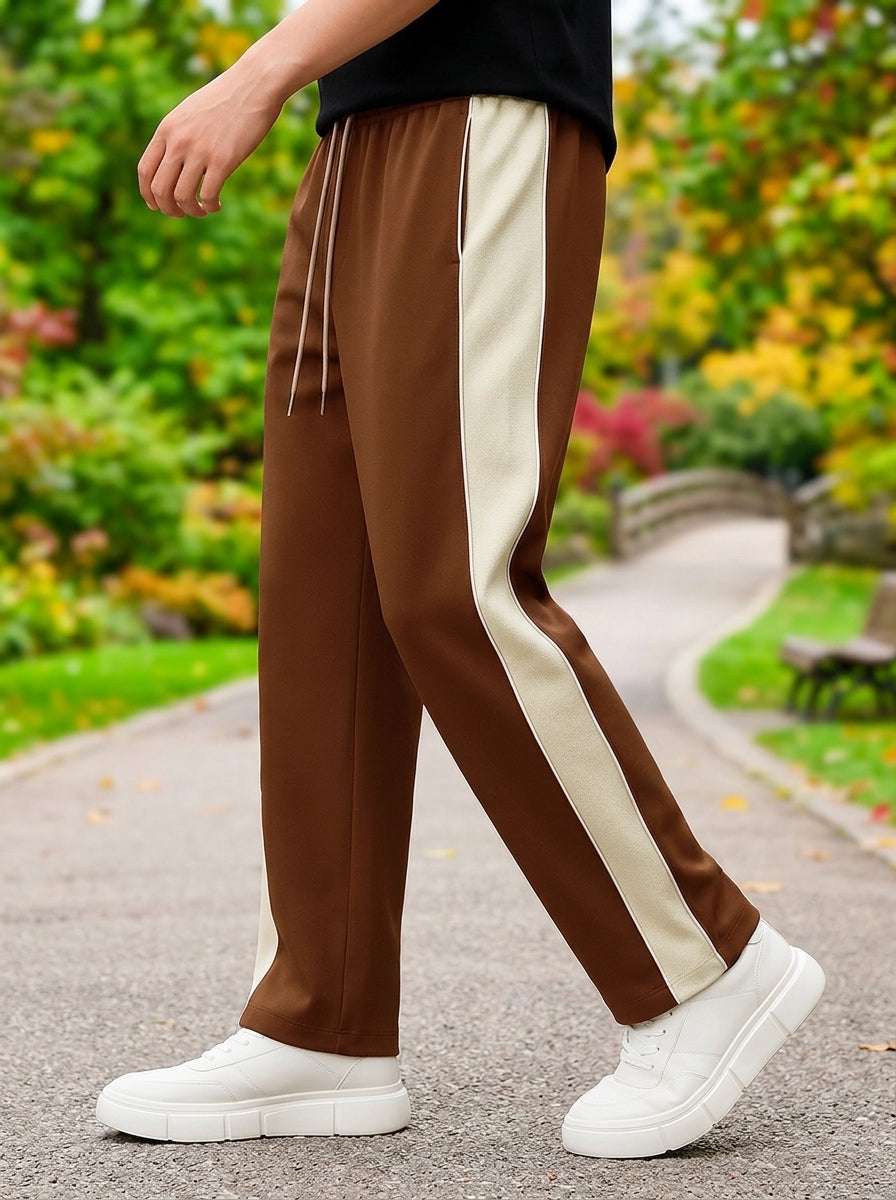 A person wearing coffee track pants with a wide cream side stripe, walking on a paved path in a garden. The pants have a drawstring waist and are paired with white sneakers.