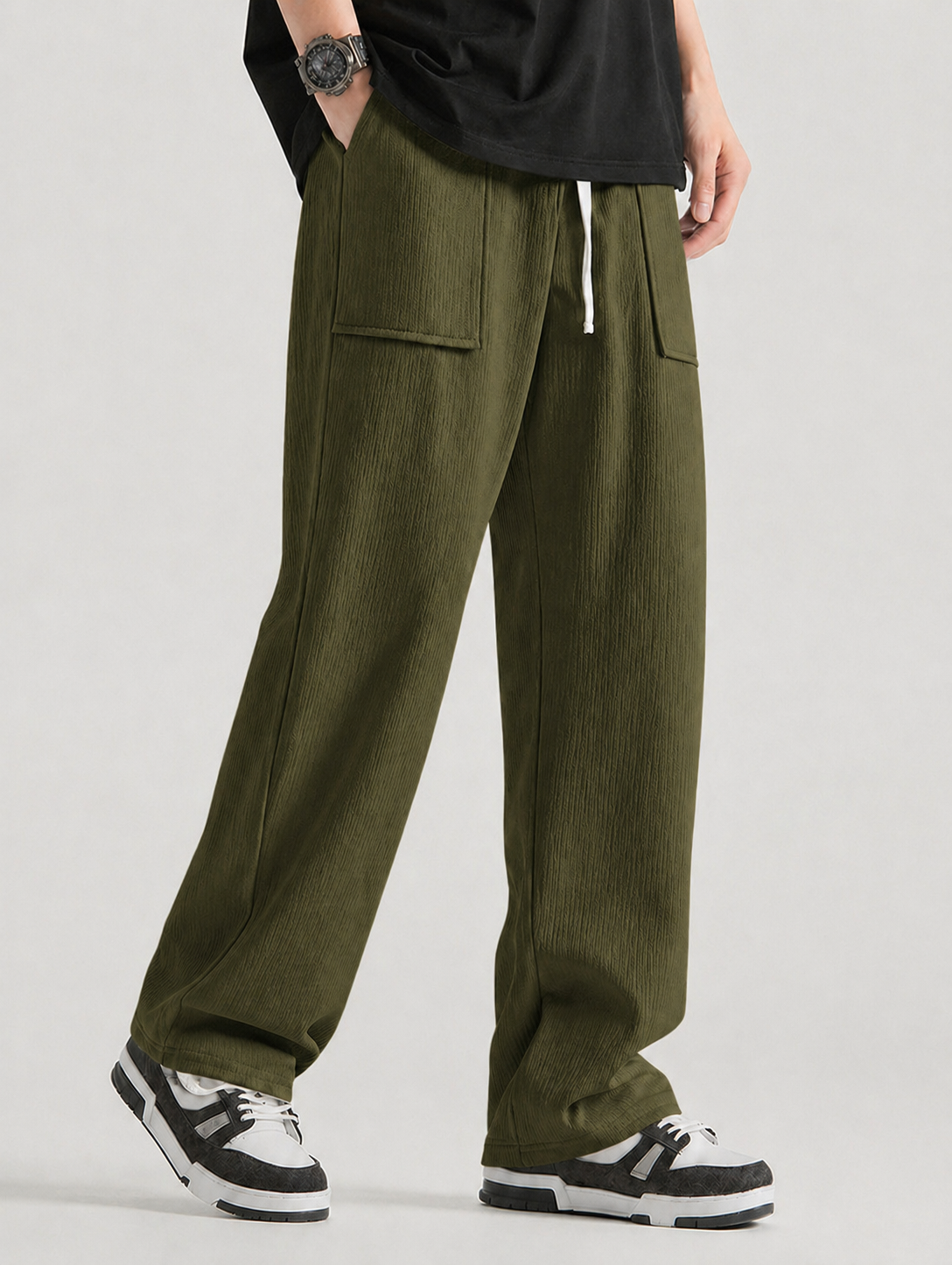 A person wearing loose olive green corduroy pants with side flap pockets, paired with a black t-shirt and black-and-white sneakers, shown from the waist down against a light gray studio background.